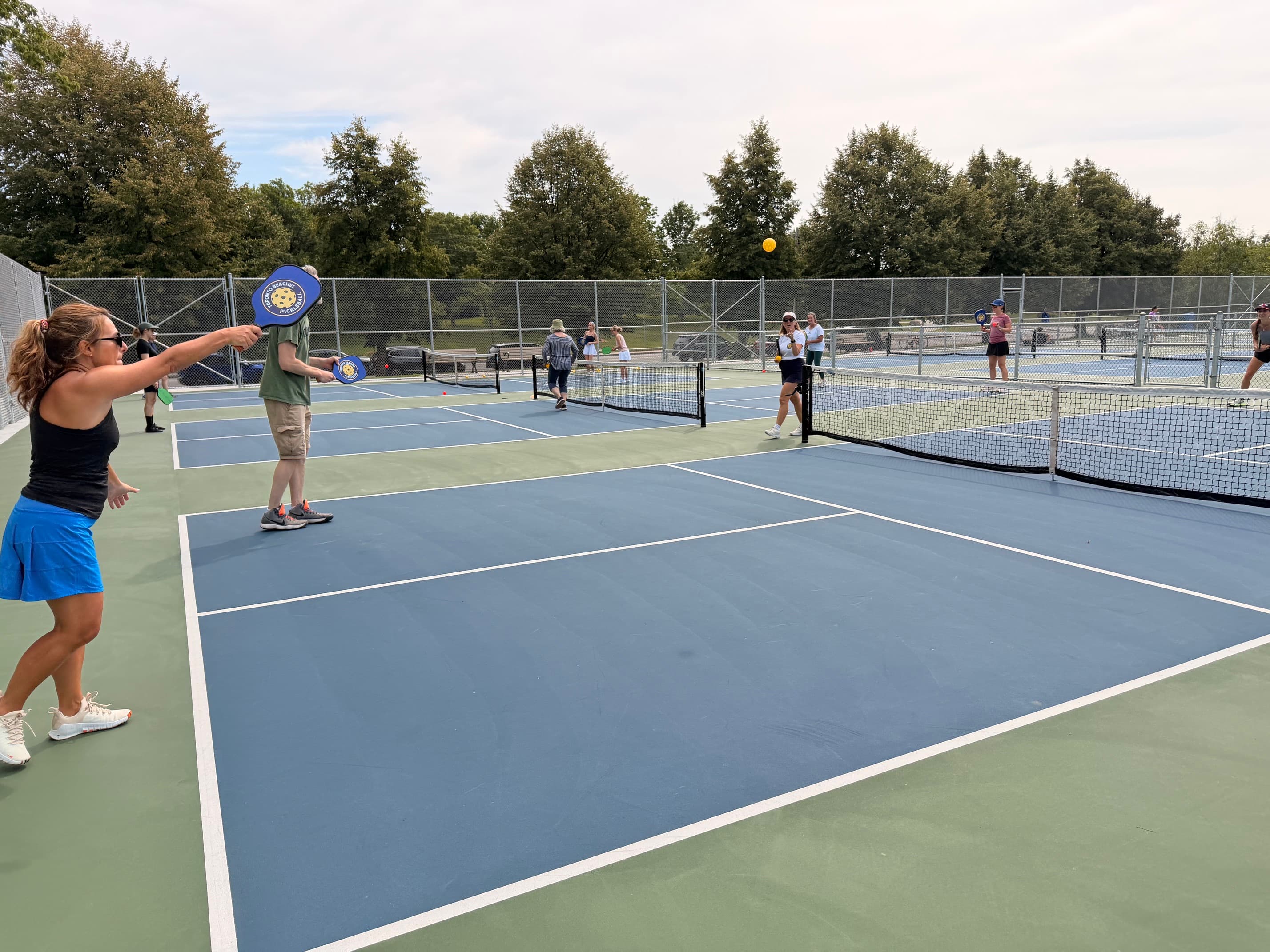 People playing pickleball on outdoor blue and green courts with a yellow ball in flight.