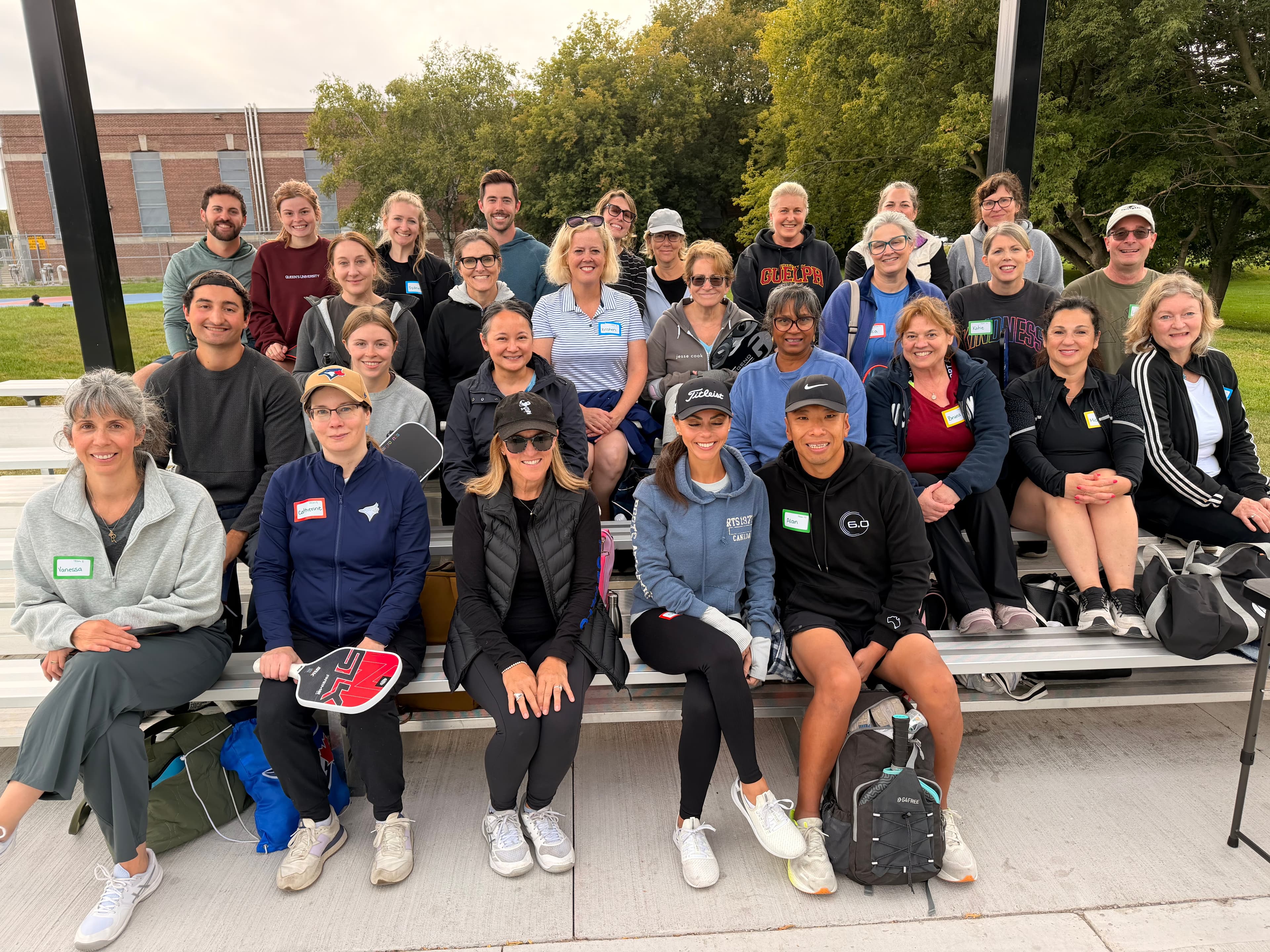 A large group of people in athletic wear pose on outdoor bleachers with pickleball paddles.