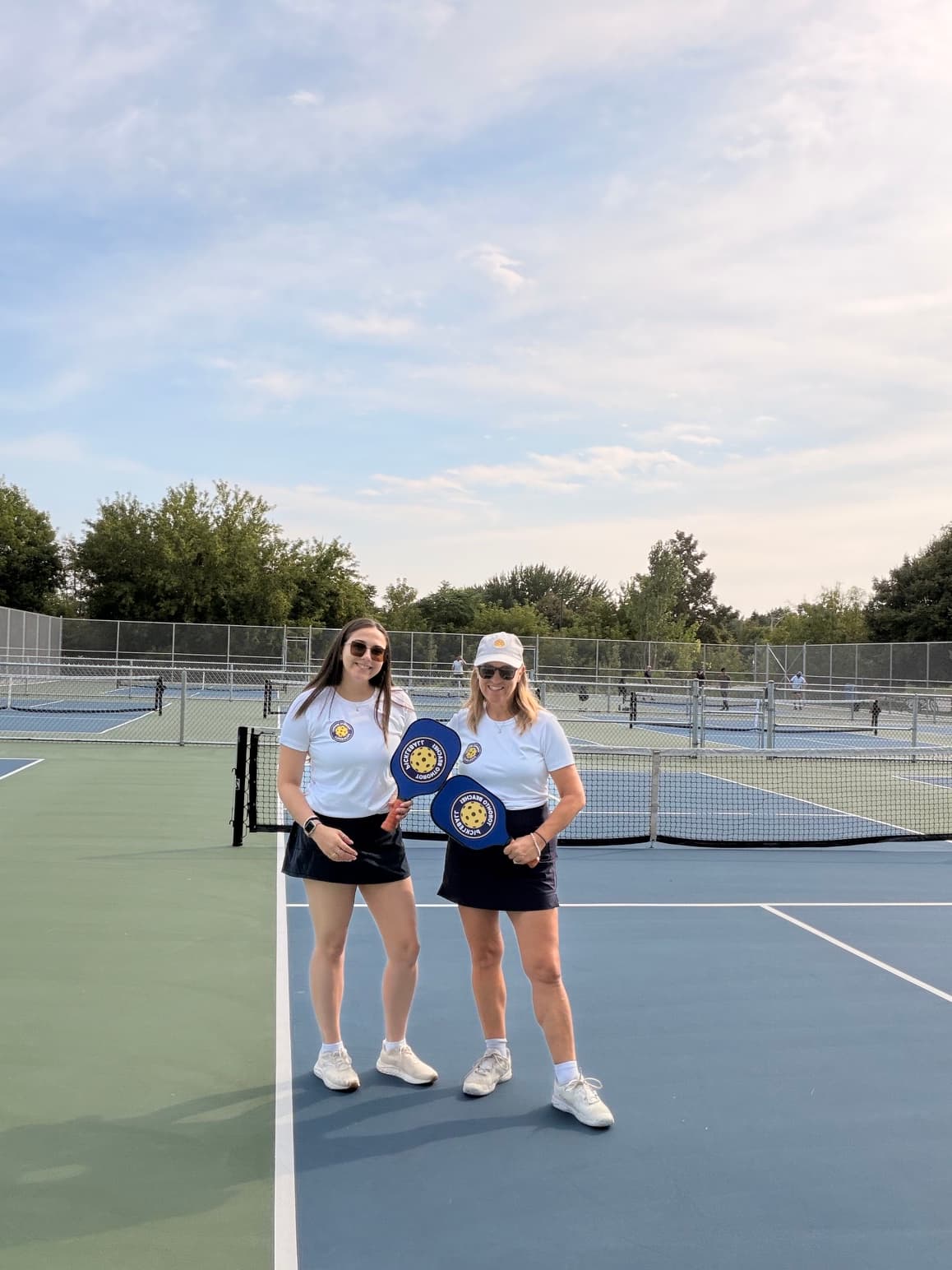 Two women in matching white shirts and black skirts hold blue pickleball paddles outdoors.