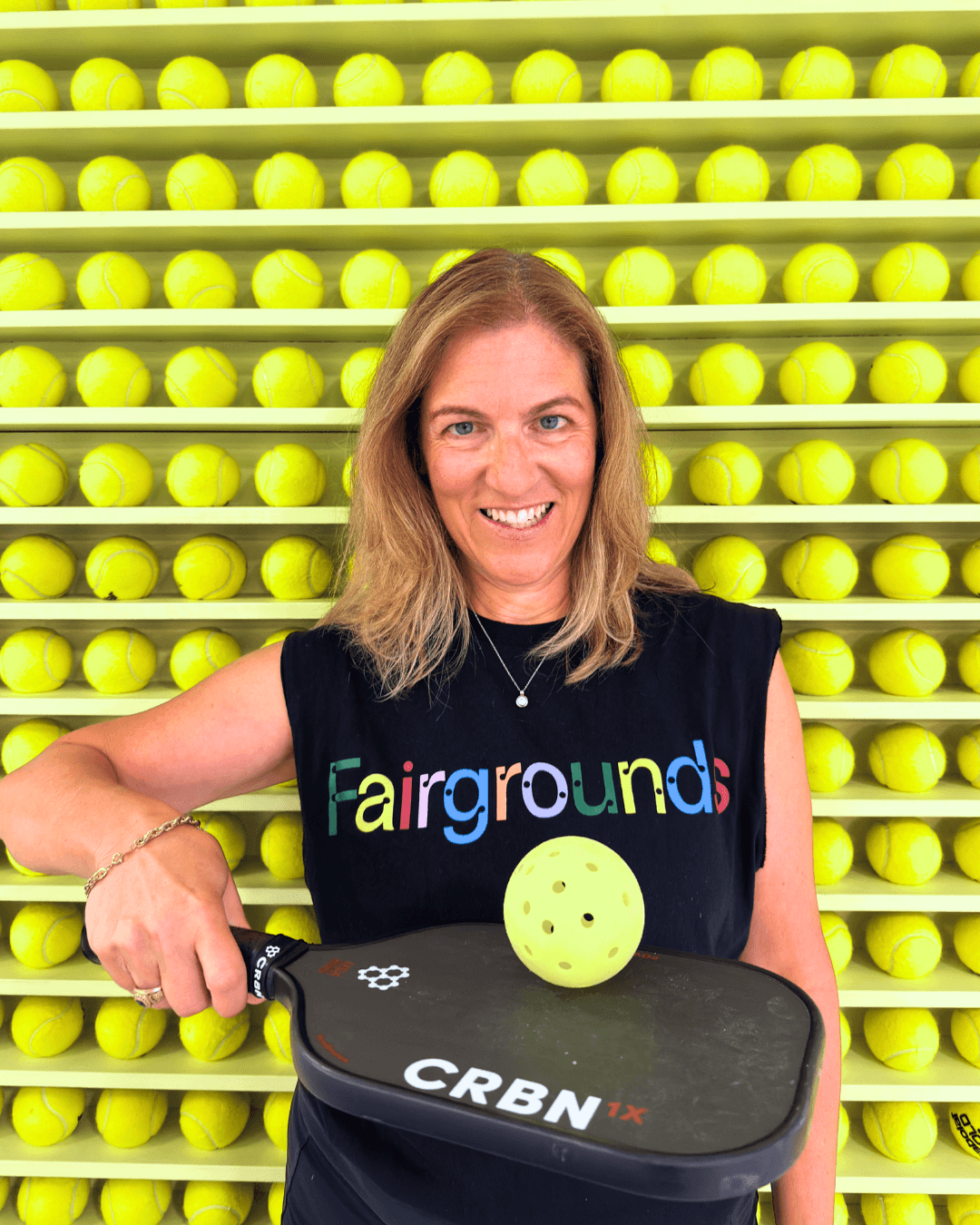 Smiling woman holding a pickleball paddle and ball against a wall of yellow tennis balls.
