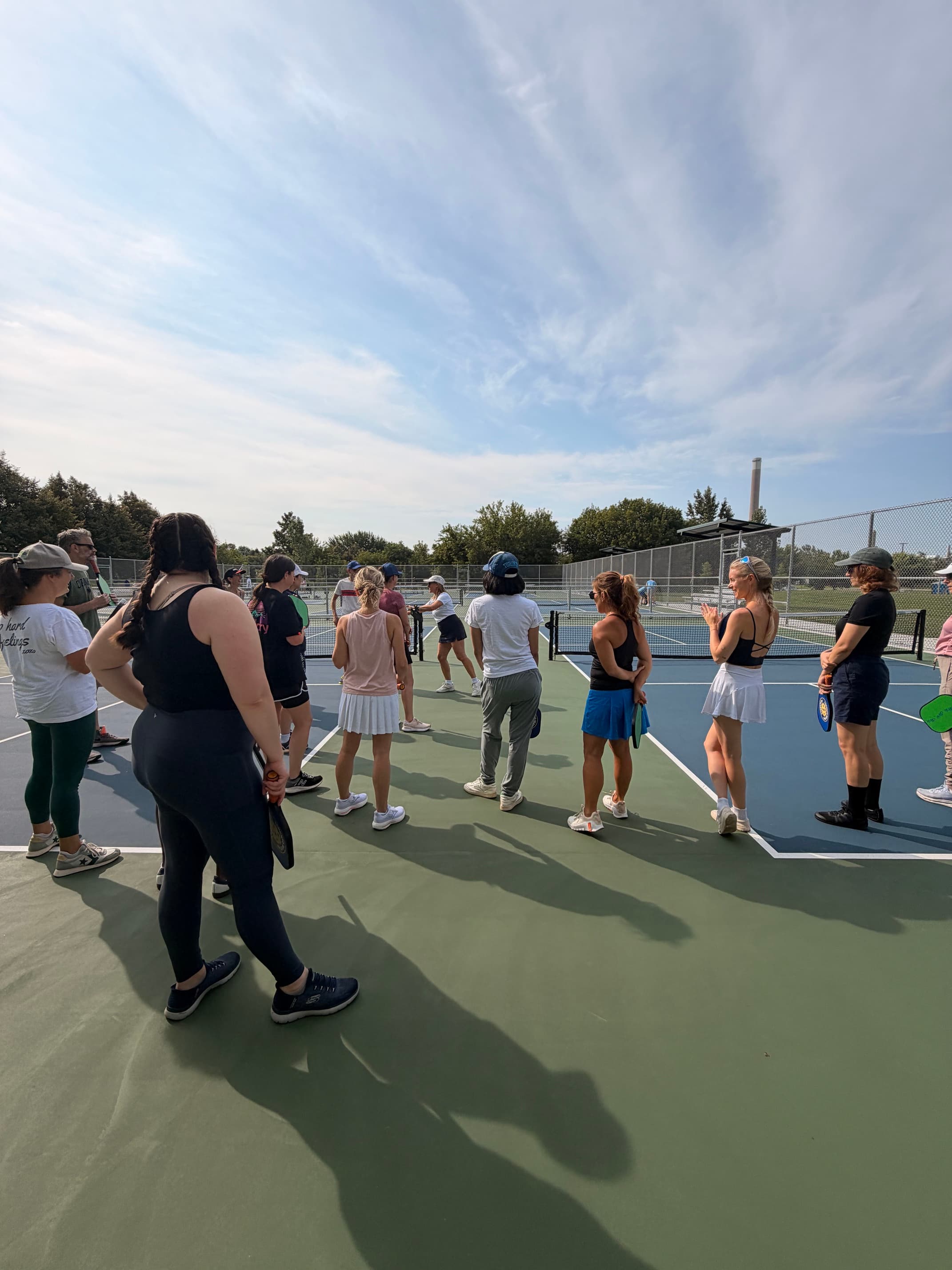 Women in athletic wear stand on a pickleball court during a sunny outdoor lesson.