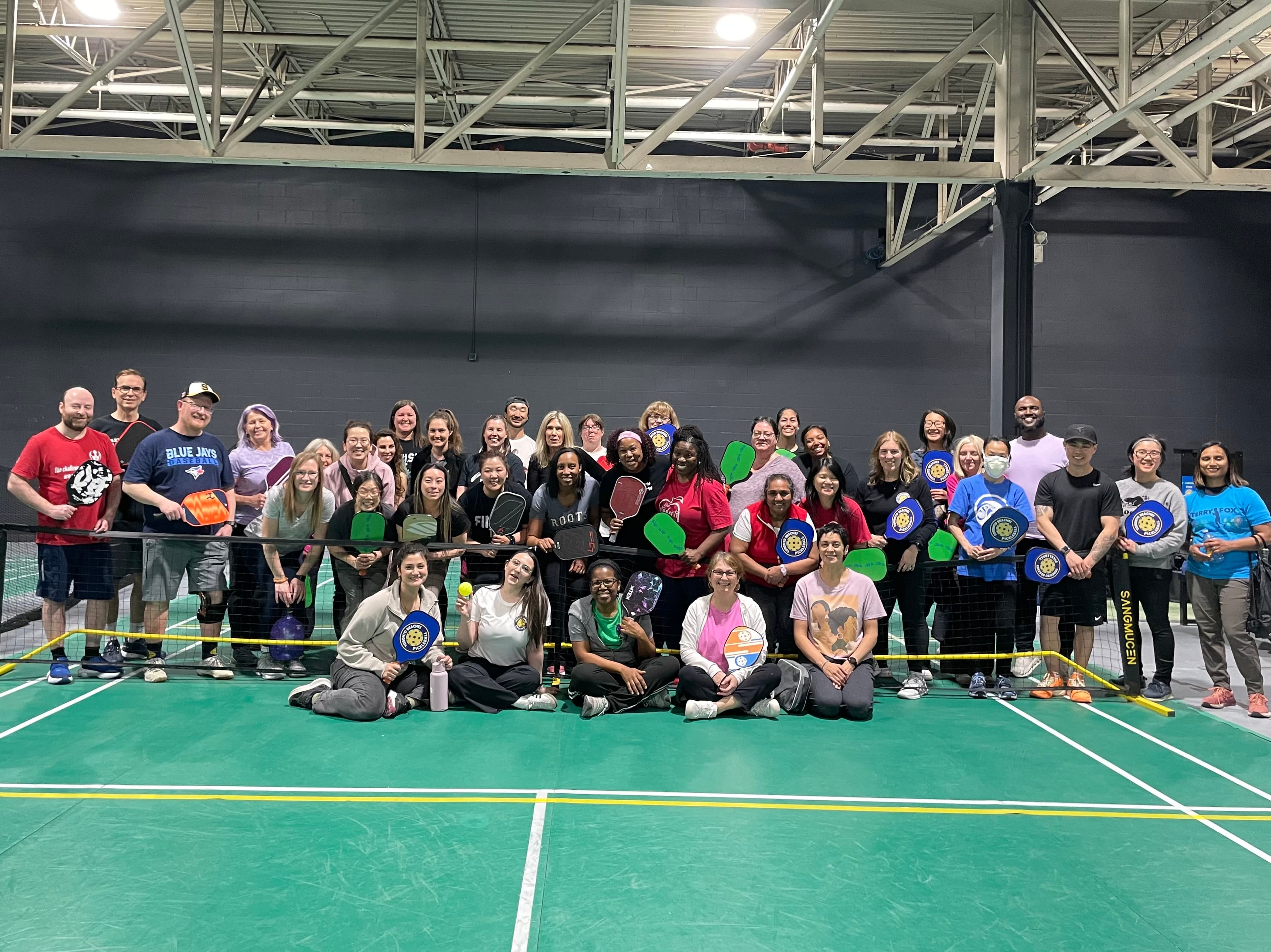 Diverse group of smiling people holding pickleball paddles on an indoor green sports court.