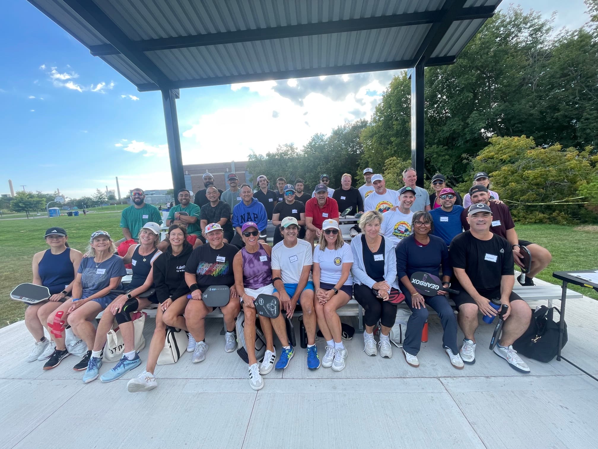 Large group of pickleball players sitting on bleachers under a pavilion, holding their paddles.