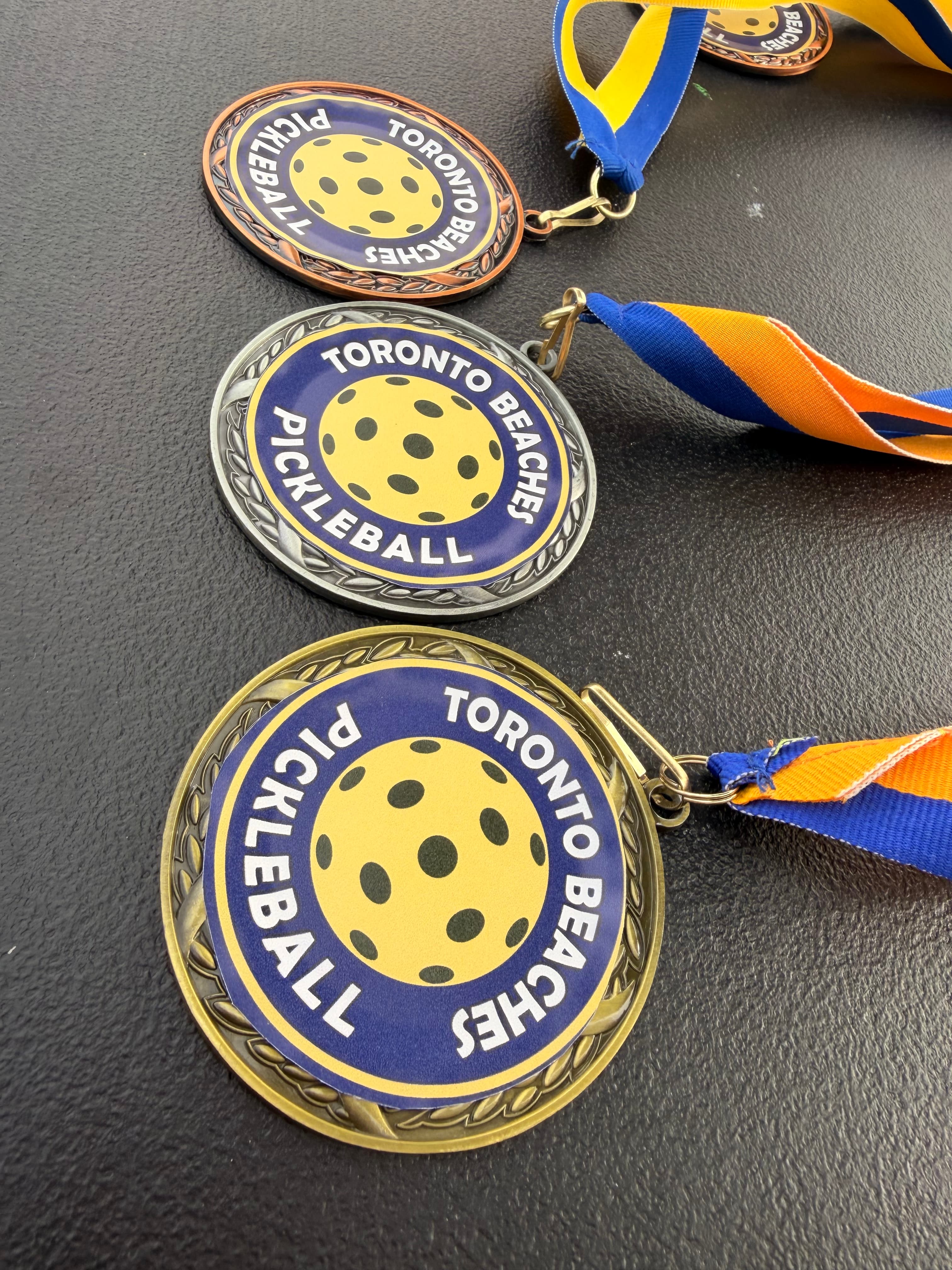 Gold, silver, and bronze Toronto Beaches Pickleball medals with blue and yellow ribbons.