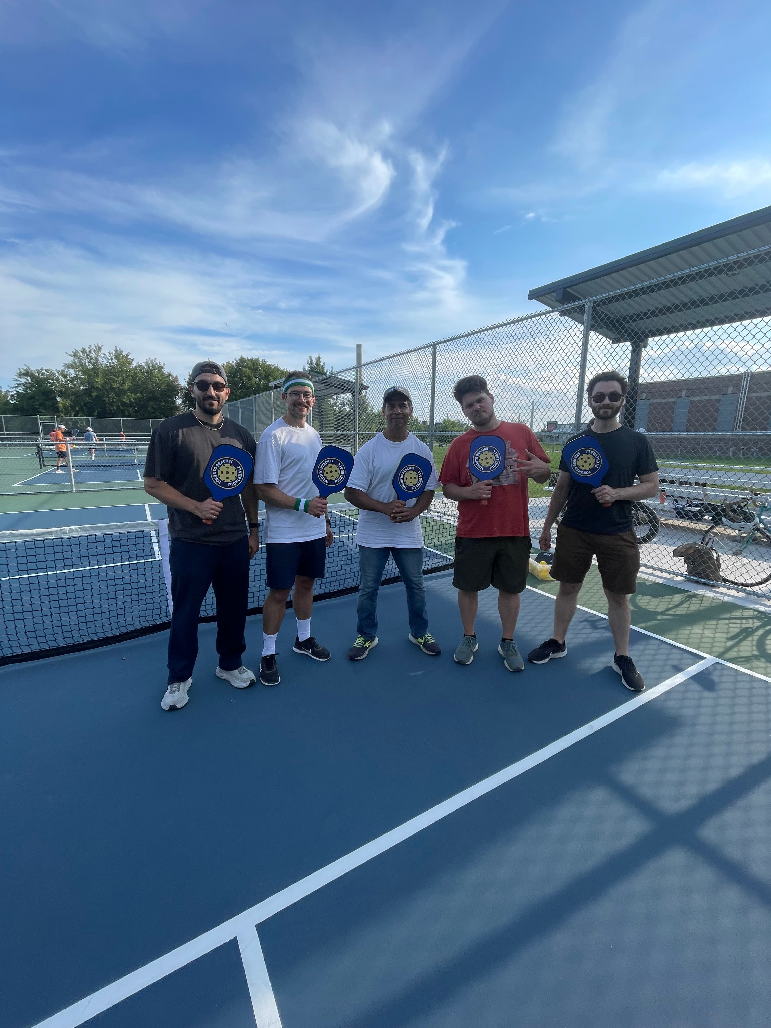 Five men pose with blue pickleball paddles on an outdoor court under a sunny sky.