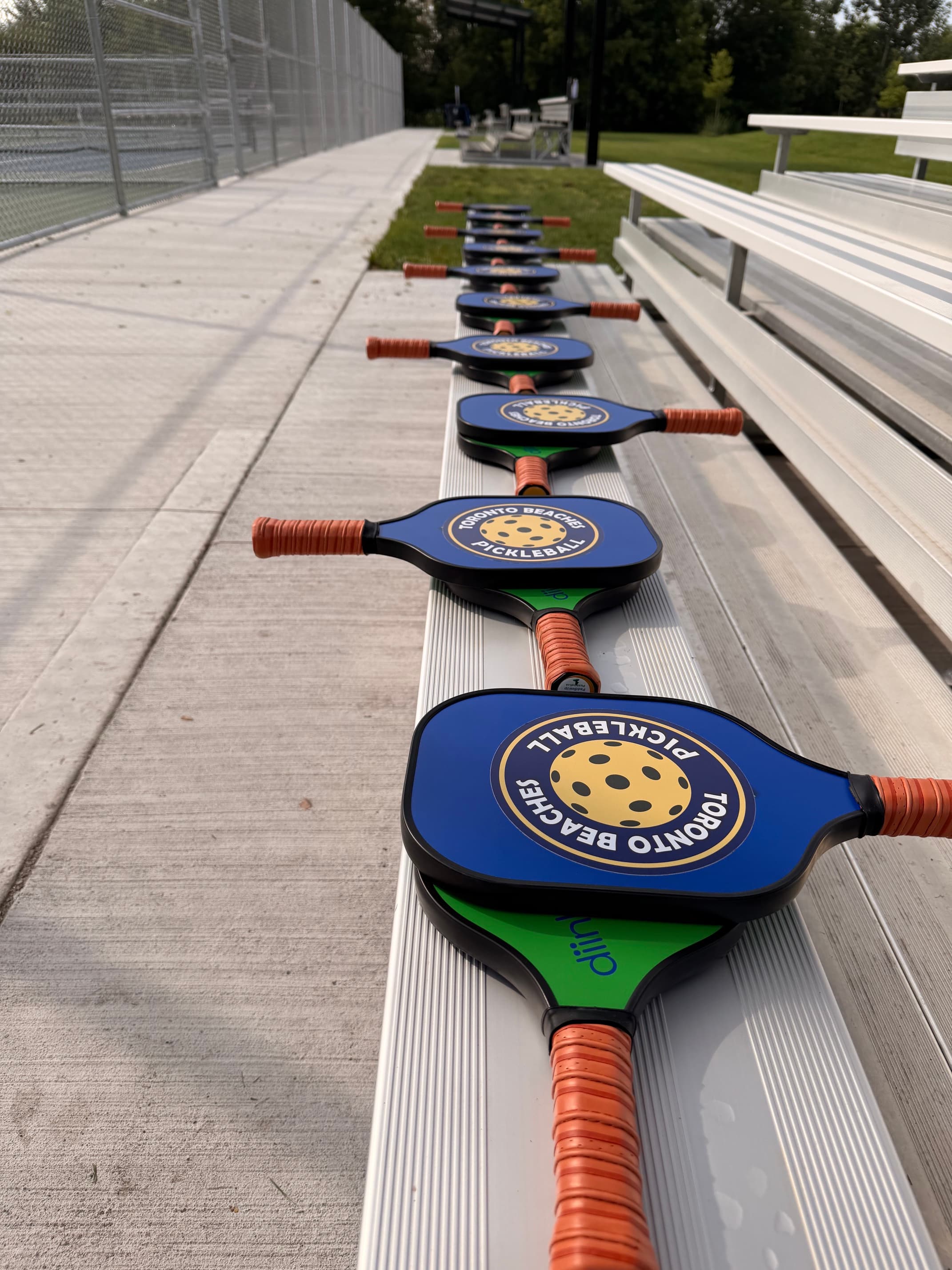 Toronto Beaches Pickleball paddles lined up on a silver metal bleacher next to a court.
