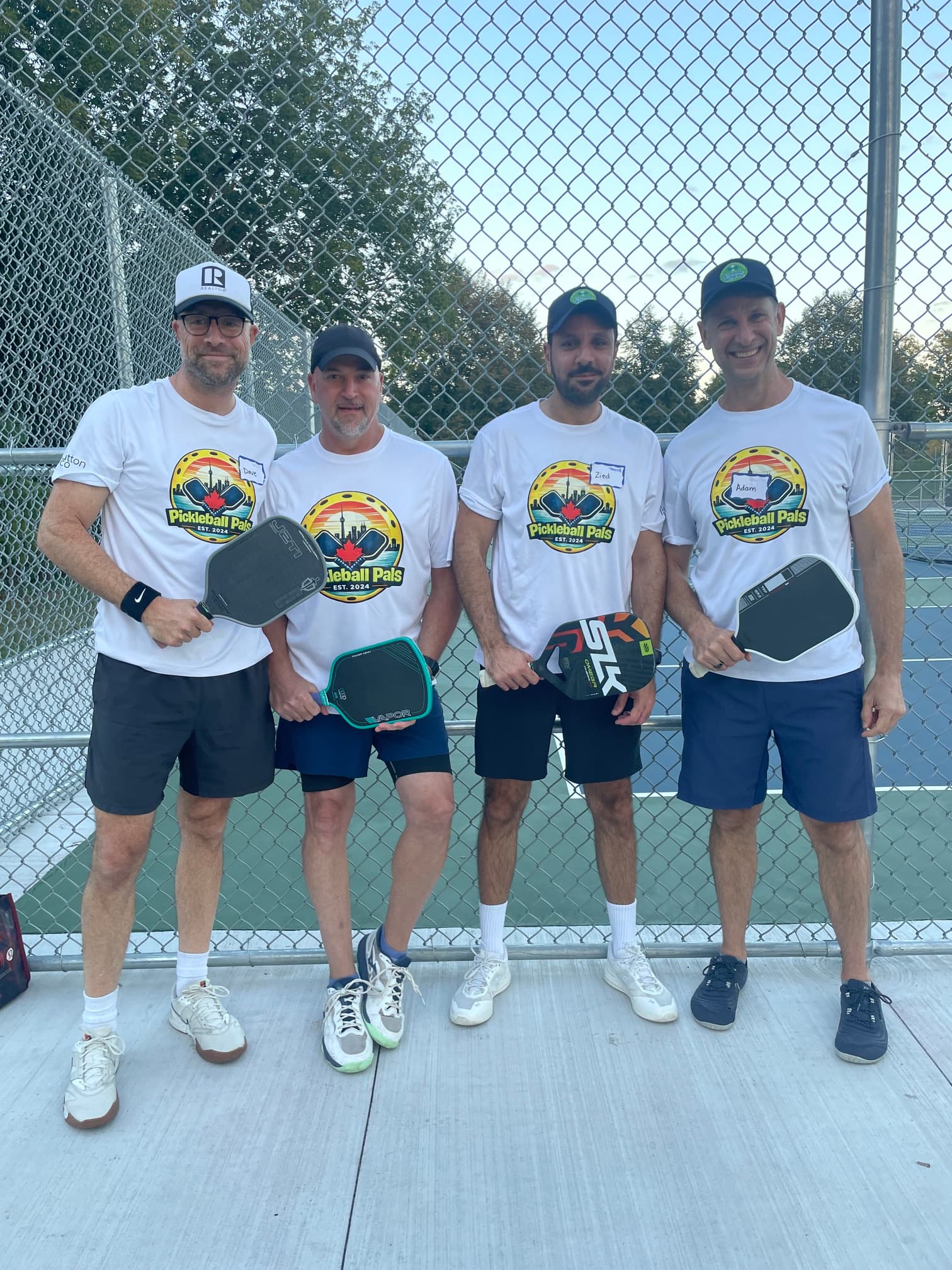 Four men in matching Pickleball Pals t-shirts hold paddles on an outdoor pickleball court.