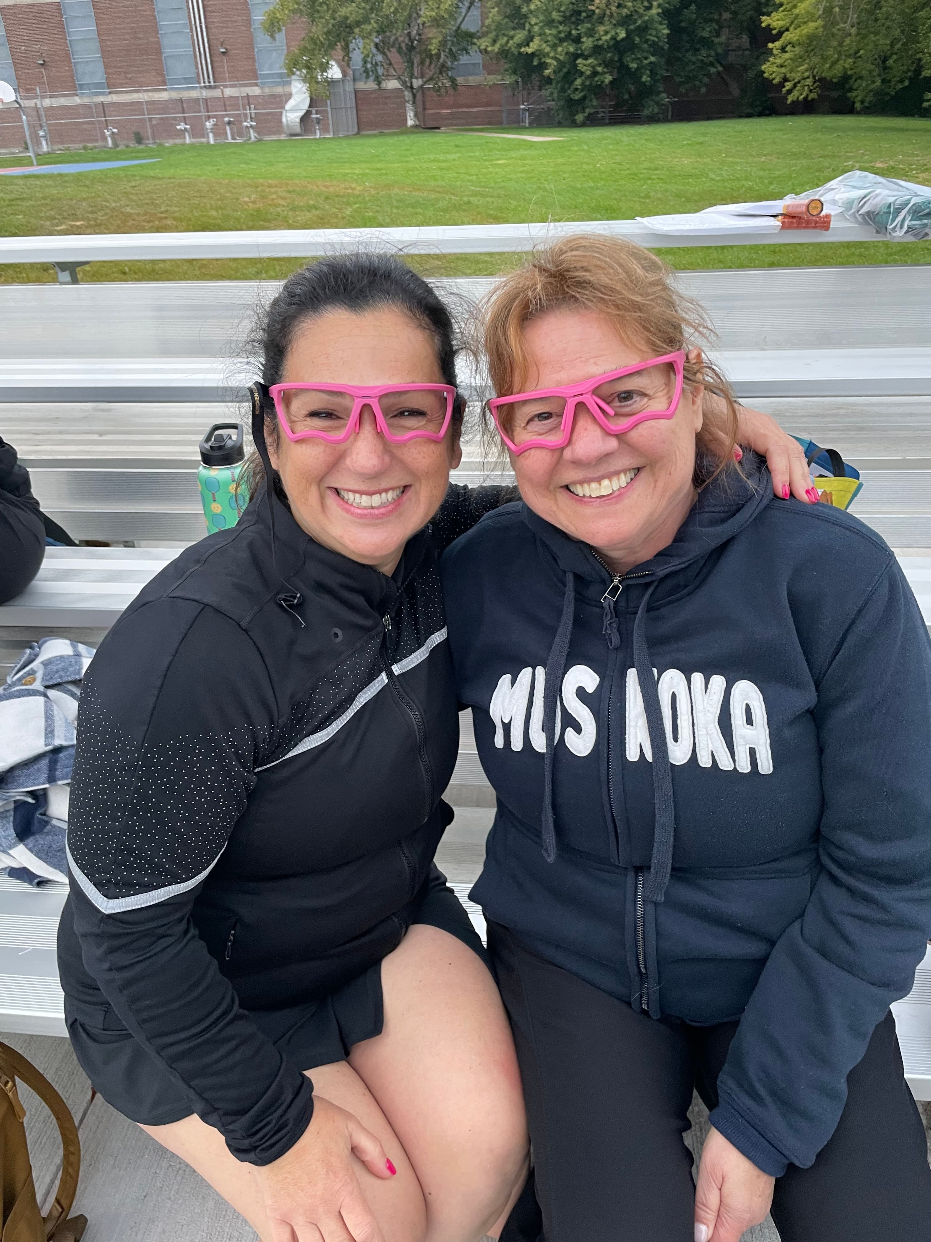 Two women sitting on bleachers, smiling and wearing matching oversized bright pink novelty glasses.