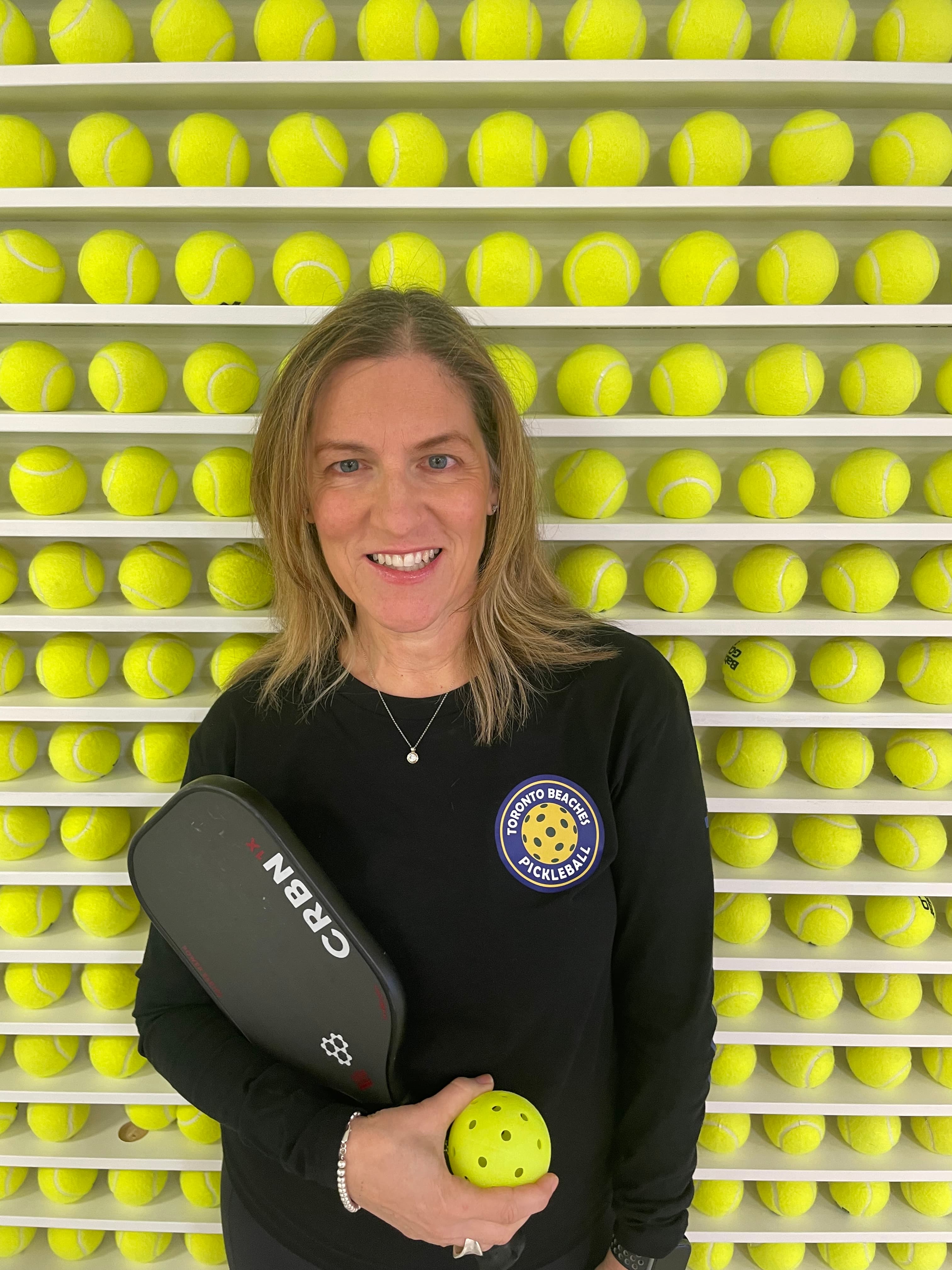Smiling woman holding a pickleball and paddle in front of a wall of tennis balls.