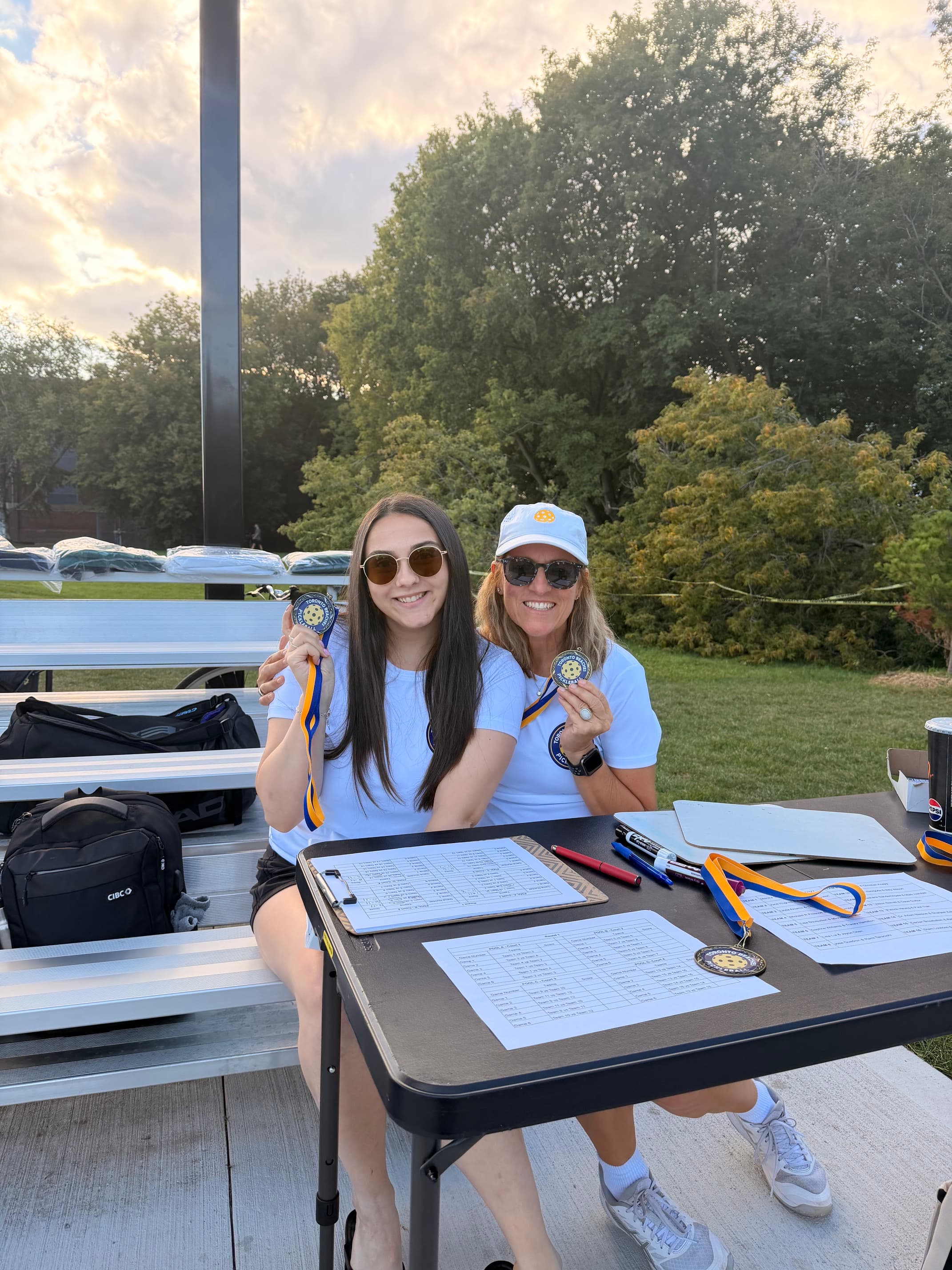Two women sit at a table outdoors, smiling and holding up pickleball tournament medals.