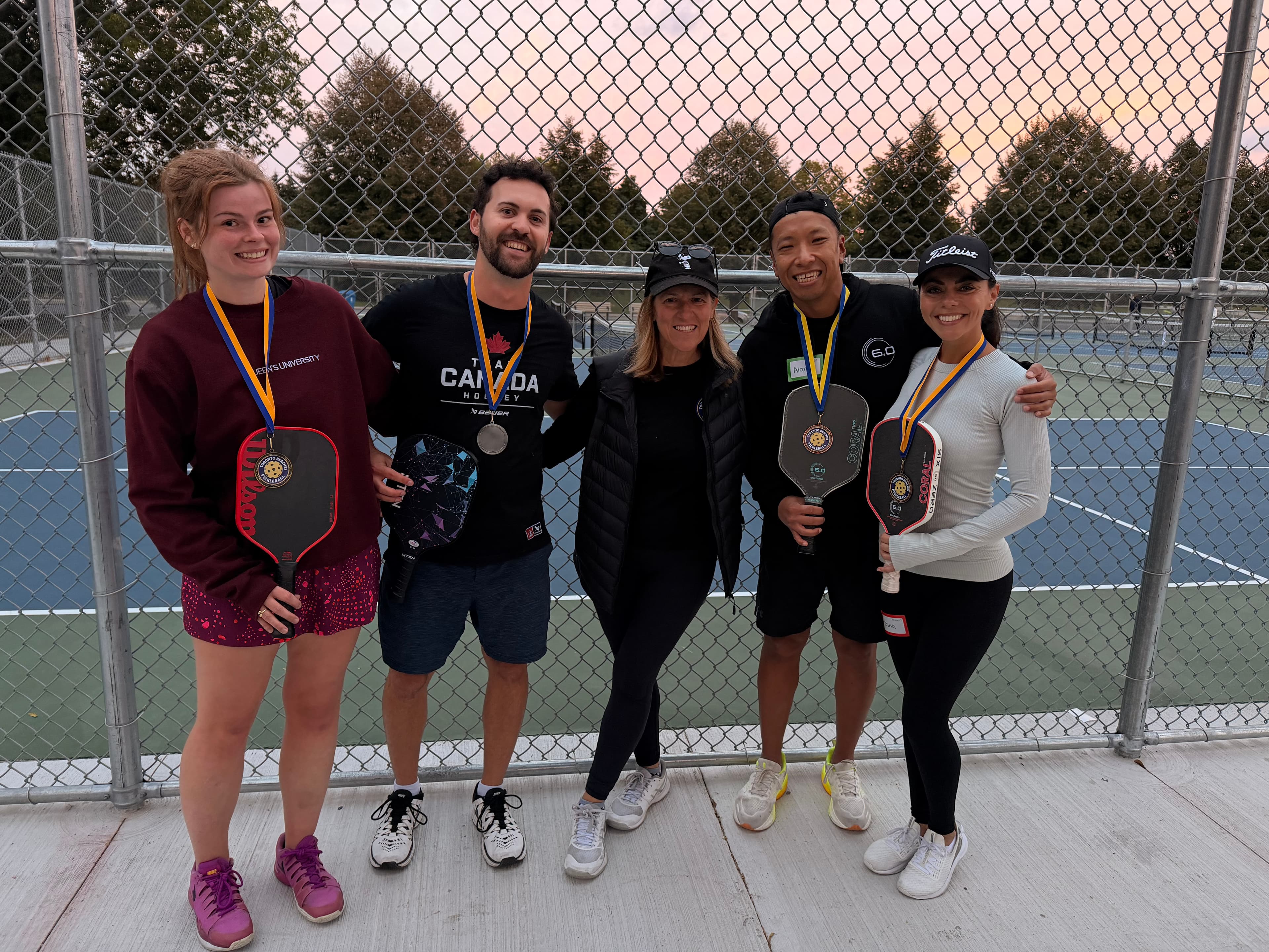 Five smiling people pose with pickleball paddles and medals on an outdoor court.